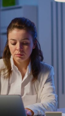 Dikey video: Yorgun kadın yönetici, dizüstü bilgisayarda yazdıktan sonra kameraya bakarak başını kaldırıyor, gece geç saatlere kadar çalışıyor. Teknoloji ağı kablosuz kullanarak odaklanmış çalışan