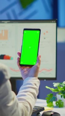 Dikey video: İş kadını gece geç saatlerde iş yerinde masasında oturan akıllı telefonun yeşil ekran monitörüne bakıyor. Masaüstü monitörünü yeşil maket, kroma tuşuyla izleyen serbest yazar