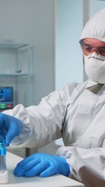 Dikey video: Steril kimya kıyafetindeki bilim adamı test tüpündeki kan örneğini analiz ediyor. Profesyonel laboratuvarda Viorolog araştırmacısı covid19 virüsüne karşı tıbbi tedavi bulmaya çalışıyor..