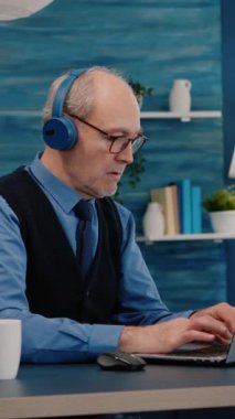 Dikey video: Kulaklıklı, müzik dinlerken evde çalışan dizüstü bilgisayardaki grafikleri kontrol eden odaklanmış eski girişimci. Emekli adam modern teknoloji kullanarak daktilo okuyor, araştırıyor.