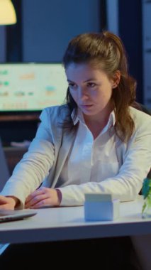 Dikey video: İş kadını dizüstü bilgisayarda yazdıktan sonra kameraya bakıp gülümsüyor ve gece geç saatlerde yeni başlayan şirkette masada oturuyor. Teknoloji ağını kablosuz kullanarak fazla mesai yapmaya odaklanmış bir çalışan