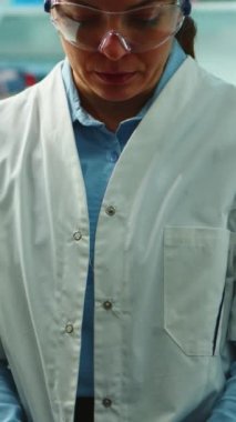 Dikey video: Gece geç saatlerde modern donanımlı laboratuvarda kameraya yorgun bakan bilim adamı hemşire görüntüsü. Uzmanlardan oluşan bir ekip araştırma için yüksek teknoloji kullanarak virüs evrimini inceliyor.