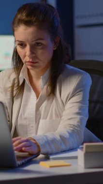 Dikey video: İş kadını projenin son teslim tarihine uymak için fazla mesai yapıyor iş ofisinde oturuyor ve gece geç saatlerde dizüstü bilgisayarla yazı yazıyor. Modern teknoloji kablosuz ağını kullanarak çalışanlarla meşgul