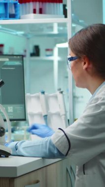 Dikey video: Mikroskop altında örneklere bakarak modern laboratuvarda yeni aşılar için gece geç saatlere kadar çalışan mikrobiyolog. Yüksek teknoloji ve kimya araçları kullanarak virüs evrimini inceleyen iş arkadaşları