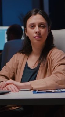 Gece geç saatlerde iş yerinde masa başında otururken kameraya bakan gülümseyen iş kadınının portresi. Pazarlama istatistiklerinde çalışan finansal istatistikleri planlayan kadın.