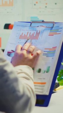 Dikey video: İş kadını elinde grafik ve mali istatistiklerle birlikte bir pano tutarken bilgisayarın önünde yeni açılan iş ofisinde otururken yakın plan. Meşgul çalışan kullanıyor