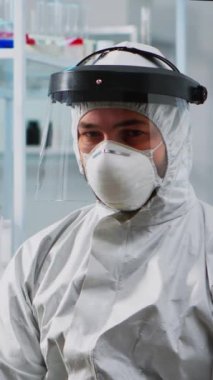 Dikey video: Donanımlı laboratuvarda kamera karşısında bitkin görünen Coperid-19 'a karşı koruyucu giysi giyen yorgun doktor. Bilimsel araştırmalar için yüksek teknoloji kullanan bilim adamları virüs evrimini inceliyor.