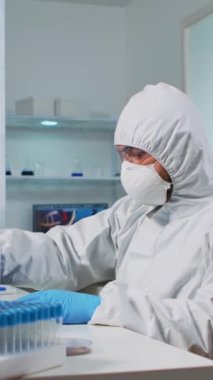 Dikey video: ppe suit içindeki biyolog sıvı örneklerini laboratuarda bilgisayarda kontrol ediyor. Ekip, yüksek teknoloji ve kimya araçları kullanarak tıbbi laboratuvarda aşı evrimini inceliyor.
