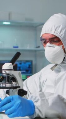 Dikey video: ppe suit giymiş bir bilim adamı laboratuvar mikroskobunun örnek aşamasına bir slayt yerleştirerek ayarlamalar yapıyor. Kimyager, çeşitli bakteriler ve doku örnekleri için çalışıyor.