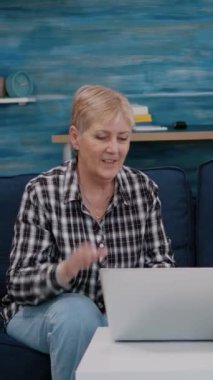 Dikey video: Koltukta otururken dizüstü bilgisayara bakıp el sallayan heyecanlı yaşlı çift. Mutlu emekli karı ve koca kanepede dinlenirken konuşarak gülüyorlar.