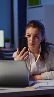 Dikey video: İş kadını, ortaklarıyla video konferansı sırasında akıllı telefon ve kulaklık kullanarak gece geç saatlerde başlangıç ofisinde tartışır. Modern teknoloji ağı çalışanı.