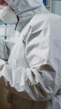 Dikey video: Koruma giysisindeki bilim adamı donanımlı laboratuarda mikroskoptan bakıyor. Virüs evrimini inceleyen kimyagerlerden oluşan bir ekip aşının geliştirilmesi için yüksek teknoloji kullanıyor.