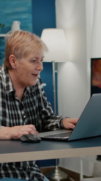 垂直ビデオ 自宅のリビングルームで働いているラップトップで良いニュースを受け取る老婦人 現代の技術読み取り入力を使用して熱狂的な先輩社員 シニア夫ながら検索 — ストック動画