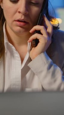 Dikey video: Gece geç saatlerde iş yerinden iş arkadaşlarıyla telefonda görüşen konsantre iş kadını. Modern teknoloji ağını kablosuz kullanarak fazla mesai yapan meşgul bir çalışan.