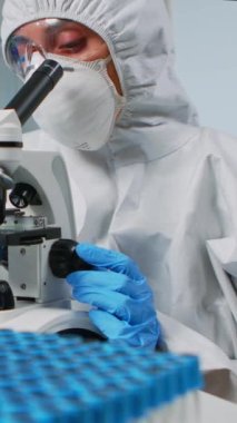 Dikey video: Pen kostümü giyen kadın araştırmacı laboratuvarda mikroskop altında örneklere bakıyor. Doktor çeşitli bakteriler, doku, kan testleri, ilaç araştırmaları ile uğraşıyor.