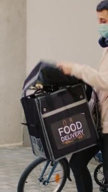 Dikey video: Yüz maskeli bir dağıtım işçisi Afrika kökenli Amerikalı müşteriye fast food siparişi veriyor, Coronavirus salgını sırasında yemek dağıtımı yapıyor. Bisikletli kadın kurye restoranda yemek dağıtıyor.