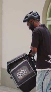 Dikey video: Restoran taşıyıcısı müşteriye fast food siparişi veriyor, ofis girişinin yakınında paket yemek dağıtıyor. Kadın müşteri, gülümseyen erkek kuryeden kese kağıdında yiyecek alıyor..