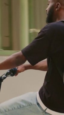 Dikey video: Paket servis sağlayıcı ofis girişine gidiyor, genç adam bisikletli yiyecek dağıtım kuryesi olarak çalışıyor. Sokakta termal çantalı ve bisiklete binen Afro-Amerikan fast food çalışanı.