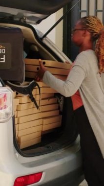 Dikey video: Yemek dağıtım elemanı müşteriye büyük pizza kutuları getiriyor, yiyecek paketlerini araç bagajından insanlara veriyor. Pizzeria kuryesi teslimat için fast food yığınları alıyor.