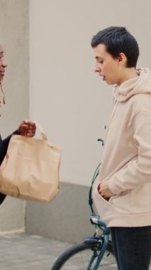 Dikey video: Afrikalı Amerikalı müşteri fast food siparişini kredi kartıyla ödüyor, bayan kurye postal terminalini tutuyor. Müşteri postacıdan yiyecek paketi almak için bağlantısız ödeme yapıyor..