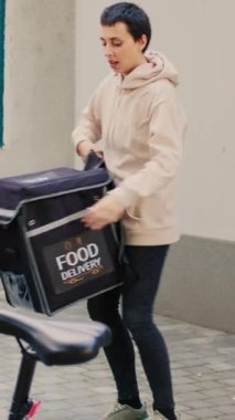 Dikey video: Paket servis elemanı girişte bekleyen müşteriye fast food siparişi veriyor, restoran paketi veriyor. Kadın teslimatçı, elinde çantasıyla öğle yemeği yiyor.