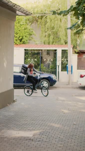 垂直视频 微笑的女人作为外卖信使和使用自行车 提供快餐餐诗意在前门 员工在阳光明媚的日子骑自行车给餐厅点菜 — 图库视频影像