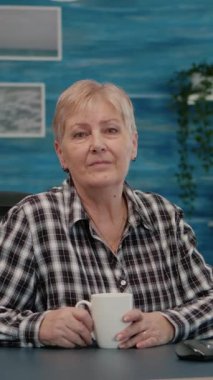 Dikey video: Yaşlı odaklı kadın iş yerinde masa başında oturmuş kameraya bakarken, yaşlı kadın arka planda televizyon seyrediyor. Emekli işadamı çevrimiçi toplantı için hazırlandı