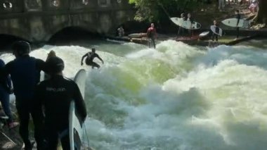 MUNICH, ALMANY - 18 Ağustos 2021: Eisbach nehrindeki bir dalgada şehir sörfçüleri. İngiliz bahçesinde nehir sörfü. 4K