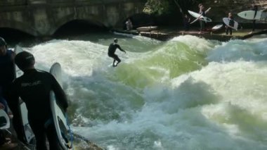 MUNICH, ALMANY - 18 Ağustos 2021: Eisbach nehrindeki bir dalgada şehir sörfçüleri. İngiliz bahçesinde nehir sörfü. 4K, Ağır çekim