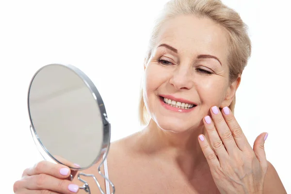 Boldog középkorú nő modell megható arc bőr tükörbe néz. Mosolygó érett idős hölgy élvezi az egészséges bőrápolás, öregedés szépség, bőrápolás kozmetikai termékek koncepció. — Stock Fotó