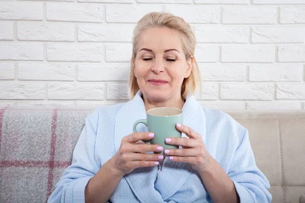 Szczęśliwa piękna kobieta w średnim wieku pije kawę na kanapie w domu — Zdjęcie stockowe