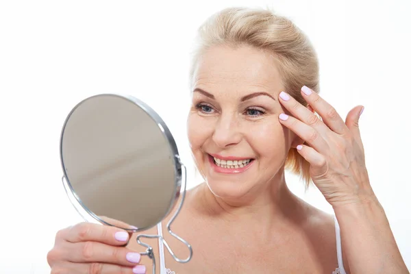 鏡を見て顔の皮膚に触れる幸せな中年女性モデル 健康な肌のケアを楽しんで成熟した老婦人を笑顔 エイジング美容 スキンケアトリートメント化粧品のコンセプト — ストック写真