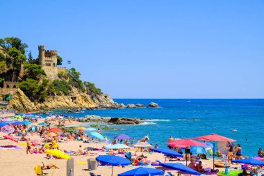 LLORET DE MAR, SPAIN - 29 Temmuz 2019: Sıcak güneşli bir günde plajdaki renkli şemsiyeler ve kayanın üzerindeki kale, Costa Brava. Plaj istirahatı kavramı. Yaz seyahati.