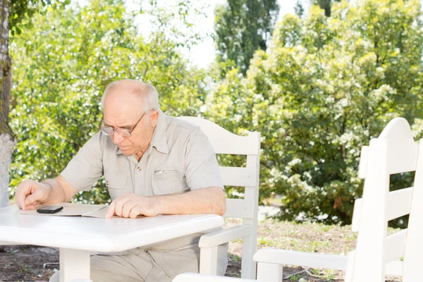 Senior man sitting reading at a table outdoors — Stock Photo, Image