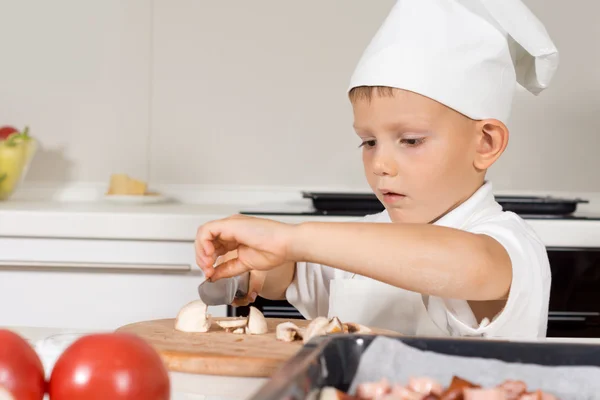 Cute little boy in a chefs toque slicing mushrooms — Stock Photo, Image