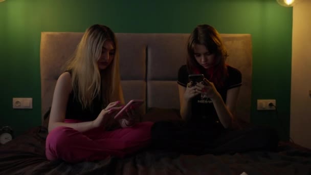 两个年轻的少女沉迷于智能手机 她们并排坐在一张昏暗的床上 在浏览自己的媒体时对彼此视而不见 — 图库视频影像