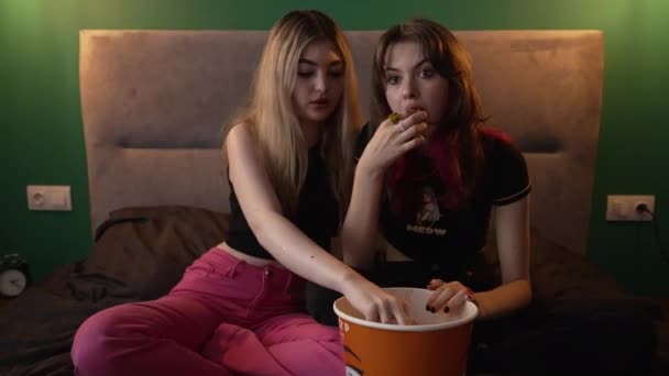 两个年轻的姐妹或朋友在昏暗的光线下一起坐在床上 享受着一盆爆米花 — 图库视频影像