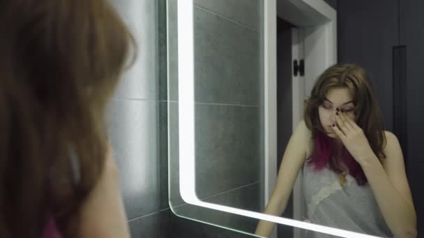 Сонная молодая женщина стоит перед зеркалом в ванной — стоковое видео