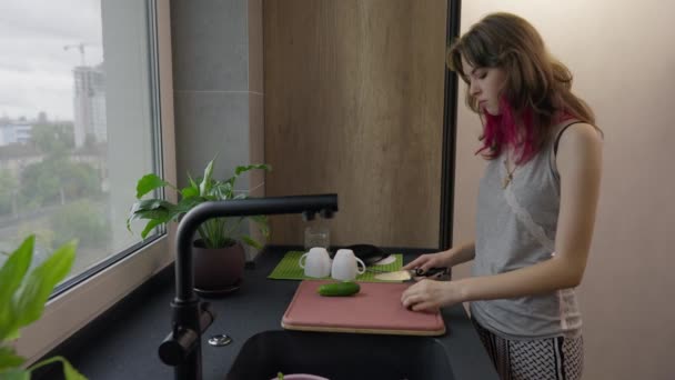 Молода жінка перекушує свіжий огірок, коли готує салат — стокове відео