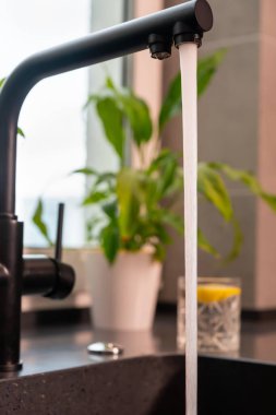 Modern bir mutfak musluğundan akan su, arka planda pencere pervazında saksı bitkisi olan lavabonun üzerinde.