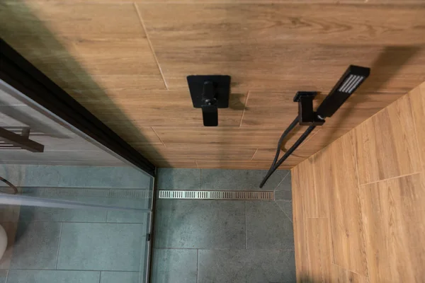 View from the top of the cubicle looking down into a shower with wood pattern tiled walls and grey tiled floor with metal drain past the mixer tap and shower head