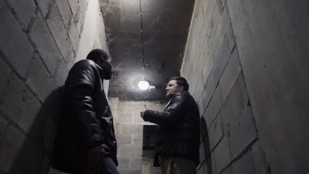 Blick auf einen Keller mit der Decke im Spinnennetz, Kaukasier und Schwarzer unterhalten sich während eines Bombardements — Stockvideo
