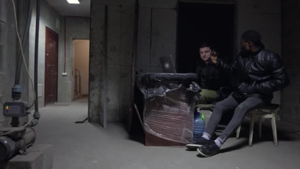 Καυκάσιος και μαύρος κάθονται σε ένα υπόγειο και συζητούν πολεμικά νέα από ένα λάπτοπ κατά τη διάρκεια βομβαρδισμού. — Αρχείο Βίντεο
