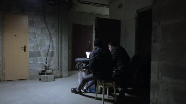 Vit kille och svart kille sitter nära öppen laptop med de senaste krigsnyheterna i en källare under ett bombardemang — Stockvideo