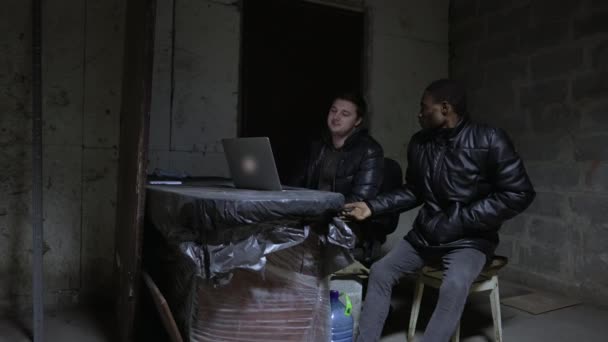 Caucasien guy et noir guy sont assis près ouvert ordinateur portable dans un sous-sol lors d'un bombardement — Video