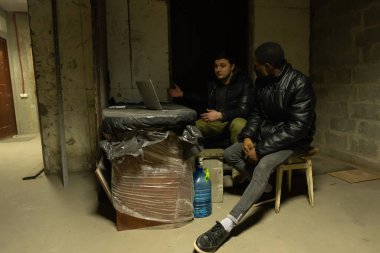 Caucasian guy and black guy are hiding in a bomb-shelter during a war and emotionally discuss the latest news, russian invasion in Ukraine clipart