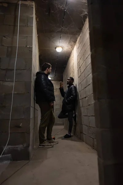 Caucasian guy and black guy are standing in a bomb-shelter during russian invasion in Ukraine