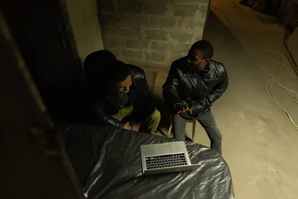 Caucasian guy and black guy are sitting near laptop in a bomb-shelter during a bombardment and having a talk, russian invasion in Ukraine