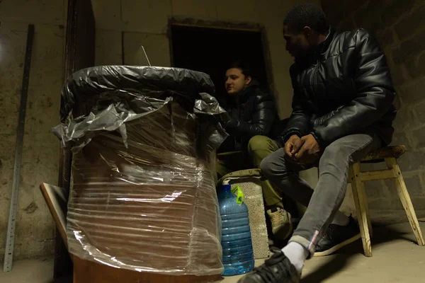 Caucasian guy and black guy are hiding in a bomb-shelter during a war and discuss the latest war news, russian invasion in Ukraine