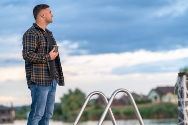 Young man standing on a boardwalk above a lake or river admiring the view wuth a cup of coffee in hand